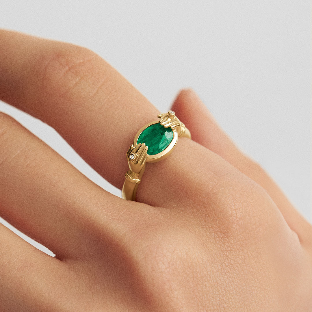 Modelo usando anel em ouro 18k de mãos adornadas com diamantes segurando esmeralda oval 